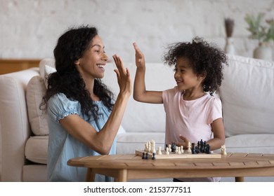 Happy Excited Black Mom Giving High Five To Cute Daughter Over Chessboard, Teaching Kid To Play Chess, Celebrating Training Success. Family Hobby, Motherhood, Mentorship Concept