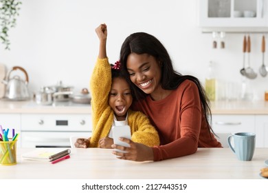 Happy Excited Black Mother And Little Daughter Celebrating Success With Smartphone While Sitting At Table In Kitchen, Cheerful African American Mom And Female Child Playing Video Games At Home