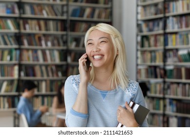 Happy Excited Blonde Asian College Student Talking On Cellphone In College Library, Laughing, Speaking, Listening With Toothy Smile, Enjoying Communication, Making Telephone Call From Public Place