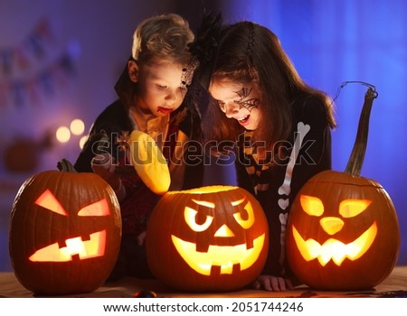 Happy excited children little boy and girl in Halloween costumes looking inside of glowing jack o lantern, carved pumpkin, while standing in dark house room decorated for all hallows eve