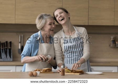 Happy excited elder mother and adult daughter woman cooking lunch together, making sandwiches at table, preparing snacks for lunch in kitchen together, laughing, chatting