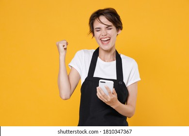 Happy Excited Fun Joyful Young Woman 20s Barista Bartender Barman Employee In White T-shirt Apron Using Mobile Cell Phone Doing Winner Gesture Isolated On Yellow Color Wall Background Studio Portrait