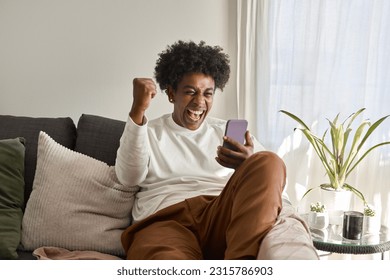 Happy excited gen z African American guy sitting on couch at home holding cellphone, celebrating online digital money betting win with yes gesture using mobile app on cell phone. Authentic shot. - Powered by Shutterstock