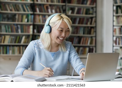 Happy Excited Gen Z Hipster College Student Girl In Headphones Enjoying Studying In Library, Consulting Teacher On Video Call, Writing Notes For Research, Class Project, Watching Webinar, Laughing