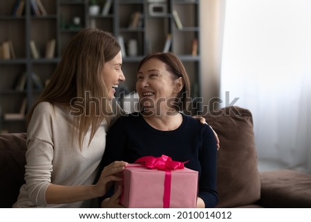 Happy excited grownup daughter giving gift to mature mom, celebrating mothers day, birthday at home, expressing love and gratitude, hugging, talking, smiling, laughing. Motherhood, family bonding