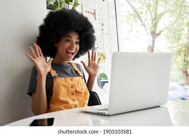 Happy excited hipster African American teen girl student with Afro hair using laptop computer video conference calling social distance friend in virtual chat, winning online sitting at table in cafe.
