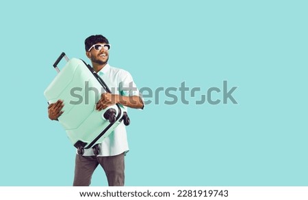 Happy excited indian guy tourist or vacationer holding travel suitcase on light blue background. Joyful ethnic young man looks towards copy space and smiles. Web banner.