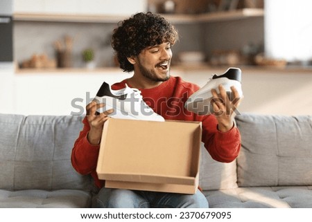 Happy excited indian man unboxing parcel with new shoes at home, smiling eastern male opening cardboard box and looking at pair of white sneakers, satisfied with online shopping, free space