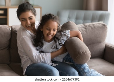 Happy excited Indian mom tickling sweet cheerful little daughter on couch, playing active pillow fighting games, smiling, laughing, enjoying motherhood, home leisure, entertainment
