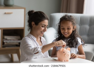 Happy excited Indian mother and kid dropping cash into piggybank. Caring mom teaching kid to save, invest money, collecting coins in piggy bank. Family savings, financial education concept
