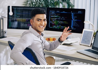 Happy Excited Indian Software Developer Sitting At Office Desk, Turning Back And Waving At Camera