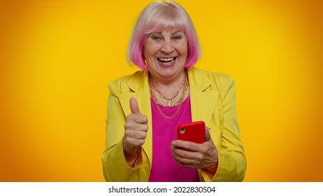 Happy Excited Joyful Elderly Granny Gray-haired Woman Use Mobile Cell Phone Typing Browsing Say Wow Yes, Found Out Great Big Win News Doing Winner Gesture. Senior Old Grandmother On Orange Background