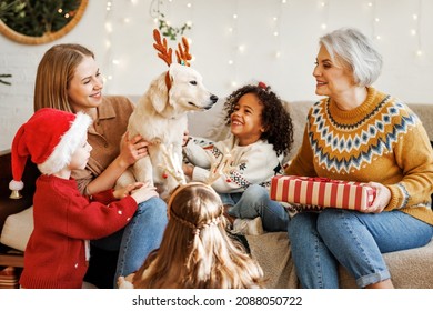 Happy Excited Kids Getting Golden Retriever Puppy In Reindeer Antlers For Christmas. Mother, Grandmother And Small Children Playing With New Friend Dog During New Year Celebration At Home