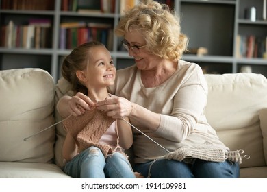 Happy excited little 6 years old cute girl enjoying learning needlework from smiling caring middle aged mature granny in eyeglasses, relaxing together on sofa at home, family hobby activity concept.