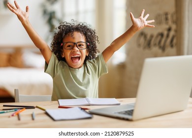 Happy excited little african american elementary school boy raising hands up and screaming with excitement during home distance education on laptop computer, kid celebrating finishing homework