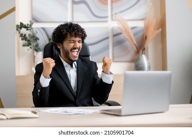 Happy excited lucky Indian man, in formal wear, business person, sits at a desk in office, got a good news, gesturing with his fists rejoicing in victory, success, good deal, big profit, is lucky man