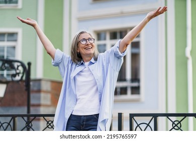 Happy Excited Mature Woman With Hands Lifted Up Wearing Glasses Standing Outdoors With Urban City Background. Senior Woman Praising God Outdoors. Giving Thanks To God Mature Woman.