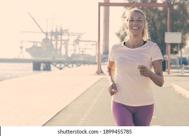 Happy Excited Mature Woman Jogging Along River Bank Outside. Senior Lady Training For Marathon. Front View. Activity And Age Concept