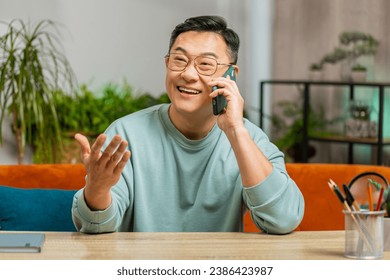 Happy excited middle aged Asian man making mobile phone call conversation with friends at home room apartment. Guy enjoying smartphone talking gossip, rumors, good news sits on couch at table desk - Powered by Shutterstock