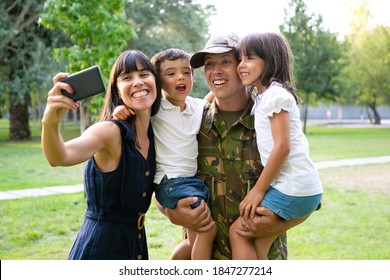 Happy Excited Military Man, His Wife And Two Kids Celebrating Dads Returning, Enjoying Leisure Time In Park, Taking Selfie On Cell Phone. Medium Shot. Family Reunion Or Returning Home Concept