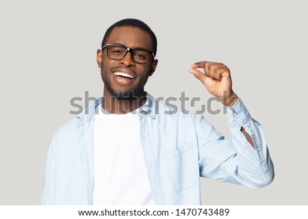 Happy excited millennial african american guy wearing eyeglasses showing with fingers small prices gesture, isolated on grey studio background. Smiling black man demonstrating little measurement.