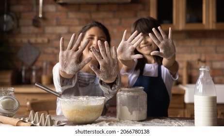 Happy excited mom and daughter baking together, showing flour hands at camera, smiling, laughing. Mother and girl cooking homemade pastry dessert for family dinner, having fun at messy kitchen table