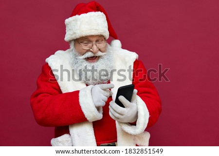 Happy excited old bearded Santa Claus wearing costume holding cell phone using mobile app on smartphone having fun, laughing, isolated on red background. Christmas promotion, xmas applications ads.
