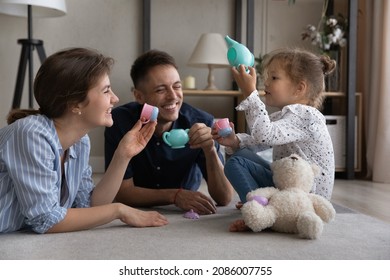 Happy Excited Parents And Cute Daughter Kid Relaxing On Warm Carpeted Floor At Home, Playing Girls Tea Party, Drinking Coffee From Toy Plastic Cups, Laughing. Parenthood, Family Playtime Activity