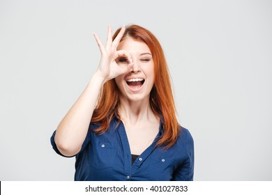 Happy excited redhead young woman looking through hole made of her fingers over white background