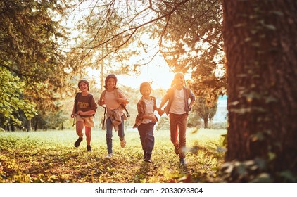 Happy Excited School Children With Backpacks In Casual Clothes Enjoying Walk In Forest On Sunny Autumn Day, Four Active Kids Boys And Girls Running And Playing Together During Camping Trip In Nature