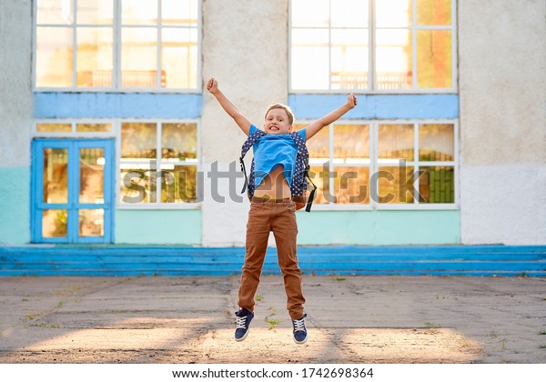 Happy excited schoolboy is happy about end of\
school year. boy with school bag, jumping for joy against\
background of school building. child is happy to start holidays.\
The concept of education