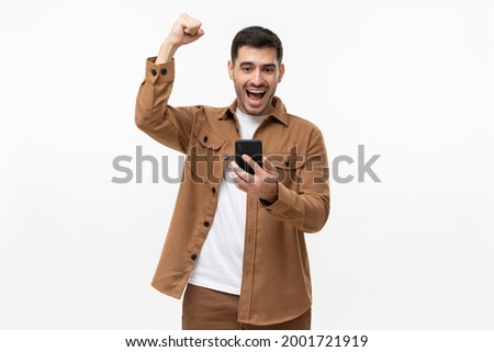Happy excited sucessful modern man holding phone and raising arm up to celebrate achievement, isolated on gray background