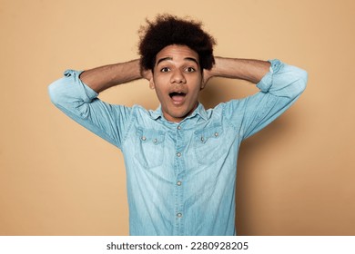Happy excited surprised african american positive youthful man in denim shirt standing against brown studio background with opened mouth and hands behind head, looking at camera with amazement - Shutterstock ID 2280928205