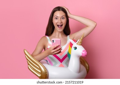 Happy excited teen girl with open mouth and inflatable unicorn ring hold phone in one hand and hold head with the other, young tik tok blogger, good news wear swimsuit isolated on pink background