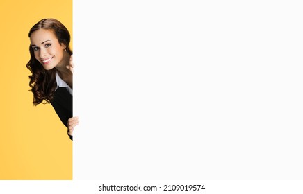 Happy excited woman in black confident suit, peek out showing blank white banner signboard with copy space for sign text. Business and ad concept. Orange yellow background. Executive employee office