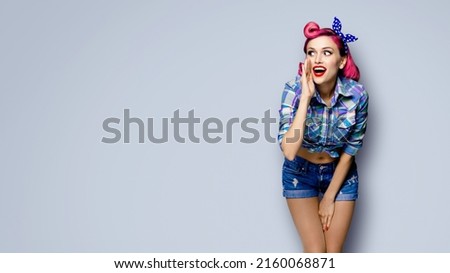 Happy excited woman holds hand near open mouth, looking aside and saying something. Girl dressed in pinup style. Red purple haired pin up model at retro fashion vintage concept, grey background.