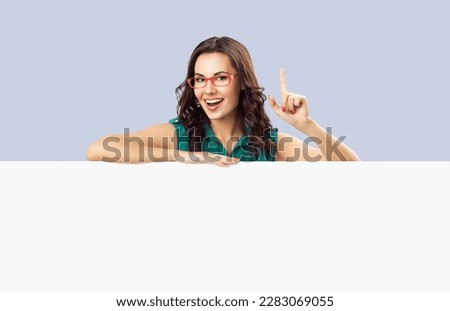 Happy excited woman with open mouth, pointing up, green cloth, red eye glasses. Mock up white bill board banner with copy space sign area, isolated on grey background. Brunette model in ad concept.