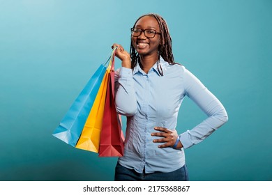 Happy excited woman smiling at camera after shopping spree at mall while holding retail store bags on blue background. Joyful young adult person holding mall purchases and enjoying goods.