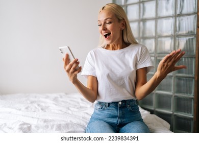 Happy excited young blonde woman getting good online news on smartphone relaxing in armchair in living room interior. Cheerful female face enjoying success using modern mobile phone at home. - Powered by Shutterstock