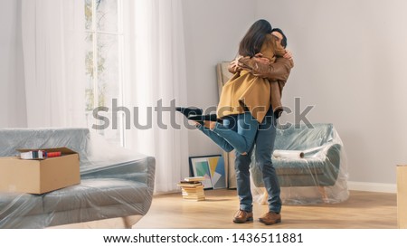 Happy and Excited Young Couple Look Around In Wonder at their Newly Purchased / Rented Apartment. Girl Jumps Into His Boyfriend's Arms Hug. Big Bright Modern Home with Cardboard Boxes Ready to Unpack.