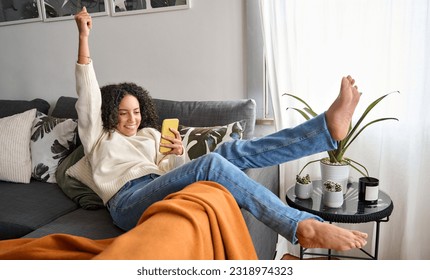 Happy excited young latin woman relaxing on couch using phone winning money in online app game. Young lucky woman feeling winner looking at cellphone, receiving great news or discount offer. - Shutterstock ID 2318974323
