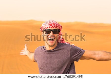 Happy and excited young male tourist standing with arms stretched against the golden Arabian desert sand dunes in the background.