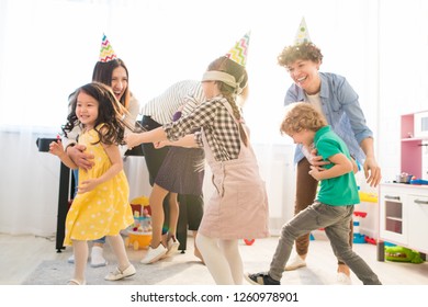 Happy Excited Young Parents In Colorful Party Hats Catching Children While Playing Blind Mans Buff With Them