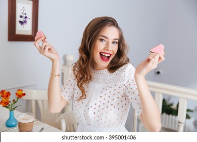 Happy excited young woman holding two heart shaped cookies sitting in cafe 