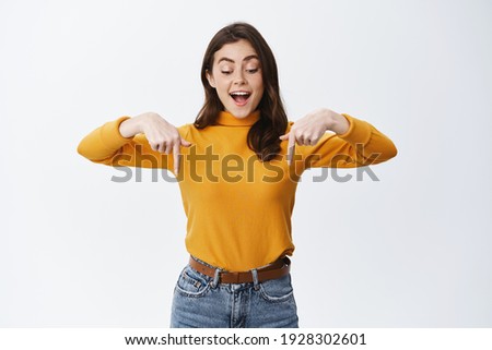 Happy and excited young woman looking mesmerized and fascinated down, pointing fingers at bottom empty space for advertisement, showing logo, white background.