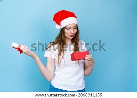 Happy excited young woman in santa claus hat with gift box on blue background in white t-shirt with surprised expression opens the box. Joy and fun and wow emotions