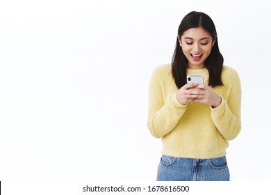 Happy Exciting Asian Girl Playing New Smartphone Game, Holding Mobile Phone, Looking At Screen With Amused Smiling Face As Watching Video Online, Texting Friend, Communicating In Internet