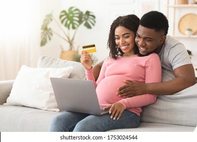 Happy expectant black couple shopping online, using laptop and credit card, sitting on couch at home, free space