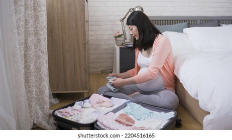 Happy Expectant Lady Sitting By A Suitcase Packed With Baby Accessories On Bedroom Floor Is Folding Clothes For Her Unborn Child.