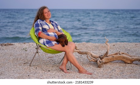 Happy expectant mother is resting in a beach chair on the seashore. curly-haired pregnant girl with her pet dog relaxes on the seashore - Shutterstock ID 2181433239